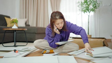 Photo for Young asian woman having problem while assembling furniture by herself. Asian woman talking on mobile phone asking how to assemble furniture. Assembling furniture concept - Royalty Free Image