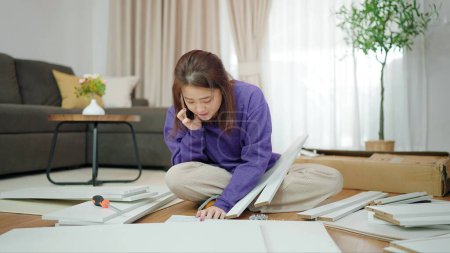 Photo for Young asian woman having problem while assembling furniture by herself. Asian woman talking on mobile phone asking how to assemble furniture. Assembling furniture concept - Royalty Free Image
