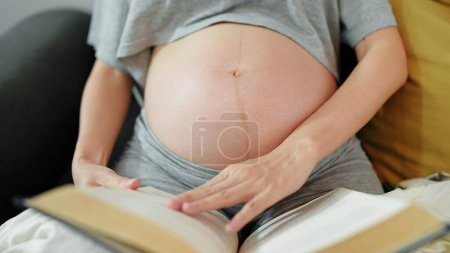 Téléchargez les photos : Femme enceinte lisant un livre assis sur un canapé confortable dans le salon à la maison. Femme enceinte relaxante profiter de la lecture d'un livre. Concept de grossesse - en image libre de droit