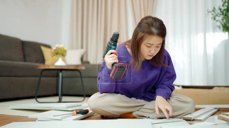 Téléchargez les photos : Les jeunes femmes asiatiques tenant une perceuse pour assembler de nouveaux meubles par elle-même à la maison. Femme assise sur le sol dans le salon tenant une perceuse pour assembler des meubles. Assemblage de meubles concept - en image libre de droit