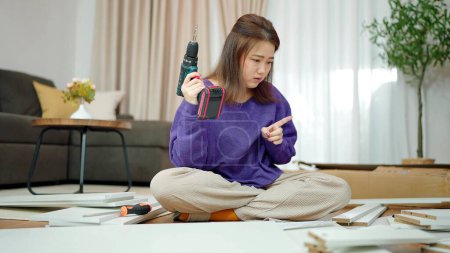Téléchargez les photos : Les jeunes femmes asiatiques tenant une perceuse pour assembler de nouveaux meubles par elle-même à la maison. Femme assise sur le sol dans le salon tenant une perceuse pour assembler des meubles. Assemblage de meubles concept - en image libre de droit