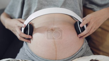 Téléchargez les photos : Femme enceinte relaxante tenant des écouteurs mettre sur l'audiothérapie du ventre bébé à naître. Femmes enceintes mettant casque jouer de la musique pour bébé dans le ventre. Concept de grossesse - en image libre de droit