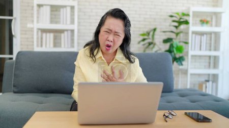 Foto de Mujer anciana asiática que sufre de dolor en el pecho de enfermedades del corazón mientras trabaja en el ordenador portátil sentado en el sofá en la sala de estar. Mujer mayor que sufre de dolor de pecho mientras está sentada en el sofá. Problema de salud - Imagen libre de derechos