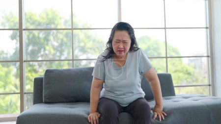 Téléchargez les photos : Femme âgée asiatique malsaine ayant des migraines ou des maux de tête alors qu'elle était assise seule dans le salon à la maison. Asiatique femme adulte souffrant de douleurs aux jambes. Concept de problème de santé - en image libre de droit