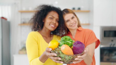 Photo for Happy lesbian woman couple holding a basket of fruits and vegetables on hands, smiling and looking at camera in kitchen room at home. Lesbian couple concept - Royalty Free Image