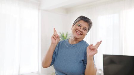 Téléchargez les photos : Gai asiatique âgés femme dans casual vêtements montrant et pointant vers le haut avec doigts numéro deux sourire à caméra à la maison - en image libre de droit