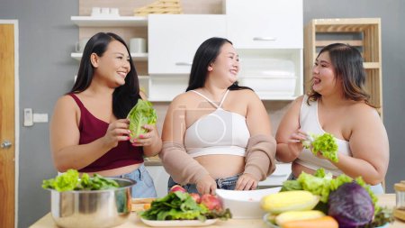 Téléchargez les photos : Groupe de jolies femmes potelées amis cuisine, préparer la salade ensemble. Trois femmes asiatiques de plus de taille parlent et cuisinent la salade dans la salle de cuisine. Concept d'alimentation saine - en image libre de droit
