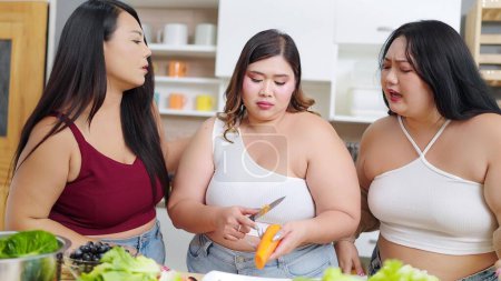Photo for Group of cheerful chubby women friends feeling sad while cooking, preparing salad together in kitchen room. Group of chubby women friends feeling sad while diet. Eating healthy food concept - Royalty Free Image