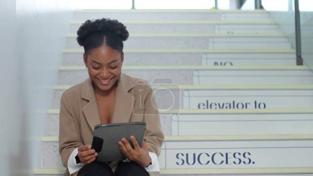 Photo for Young Africa America businesswoman using a digital tablet while sitting on the stairs at office. Businesswoman enjoy browsing on tablet while taking a break - Royalty Free Image