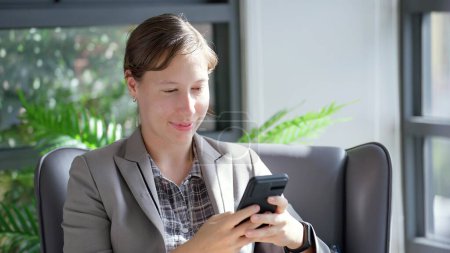 Téléchargez les photos : Jeune femme d'affaires caucasienne tapant sur smartphone alors qu'elle était assise au bureau. Bonne femme d'affaires bavarder avec un ami via smartphone sur les médias sociaux - en image libre de droit
