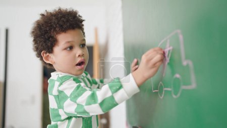 Téléchargez les photos : Afro-Américain mignon petit garçon écrit sur un tableau à craie avec de la craie en classe à l'école. Le petit garçon écrit sur un tableau vert en classe. Retour à l'école. Concept d'éducation - en image libre de droit