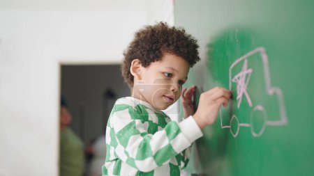 Photo for African American cute little child boy writes on a chalk board with chalk in classroom at school. Little boy writes on green board in classroom. Back to School. Education concept - Royalty Free Image