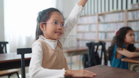 Téléchargez les photos : Active excellente écolière asiatique dans des lunettes levant la main répondent aux questions en classe à l'école. Portrait d'enfants qui étudient en classe à l'école. Concept d'apprentissage des enfants - en image libre de droit