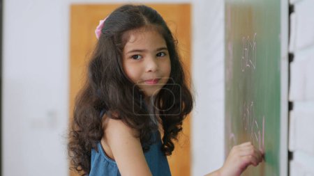 Photo for Asian little girl pupil writing or drawing on chalkboard smiling and looking at camera. Little schoolgirl smiling and looking at camera. Education and learning concept - Royalty Free Image