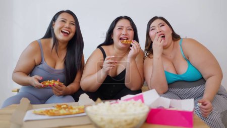 Foto de Grupo de mujeres amigas asiáticas con sobrepeso disfrutan comiendo comida poco saludable mientras ven televisión juntas en casa. Comida chatarra, Comida rápida, Concepto de comida no saludable - Imagen libre de derechos