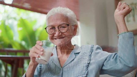 Téléchargez les photos : Les personnes âgées asiatiques attractives boivent un verre de lait pendant leurs loisirs à la maison. Femme âgée buvant du lait frais le matin pour les soins de santé. Personnes âgées mode de vie sain - en image libre de droit