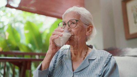 Téléchargez les photos : Les personnes âgées asiatiques attractives boivent un verre de lait pendant leurs loisirs à la maison. Femme âgée buvant du lait frais le matin pour les soins de santé. Personnes âgées mode de vie sain - en image libre de droit