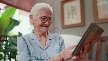 Téléchargez les photos : Heureuse femme asiatique âgée regardant à travers un album photo de famille tout temps libre à la maison. Grand-mère aînée âgée regardant des photos de famille. Moments importants et mémoire de la vie - en image libre de droit