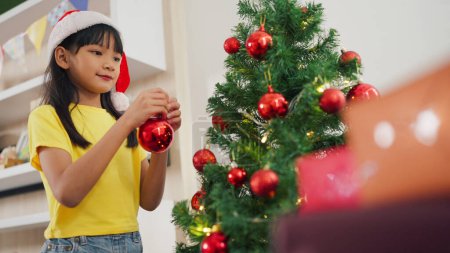 Photo for Happy asian little girl decorating the Christmas tree at home. A girl hanging decorative toy balls on Christmas tree branch. Merry Christmas and Happy Holiday. Christmas tree - Royalty Free Image