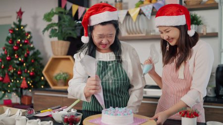 Téléchargez les photos : Joyeux asiatique mère âgée et jeune fille aider à presser la crème sur un gâteau ensemble. Mère et fille font des gâteaux pour Noël. Gâteau décoré. Vacances festives - en image libre de droit