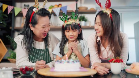 Photo for Asian little girl make a wish and blowing candles on cake with grandmother and mother in kitchen room at home. Family celebrating at home.Cheerful little girl celebrate birthday party with her parents - Royalty Free Image