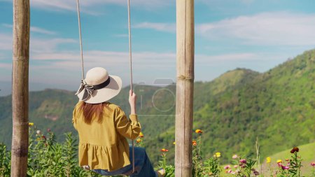 Téléchargez les photos : Vue arrière de jeune touriste asiatique assise balançant sur une balançoire belle vue sur le ciel et la montagne par temps ensoleillé. Vacances d'été. Vie heureuse et liberté. Concept de voyage - en image libre de droit
