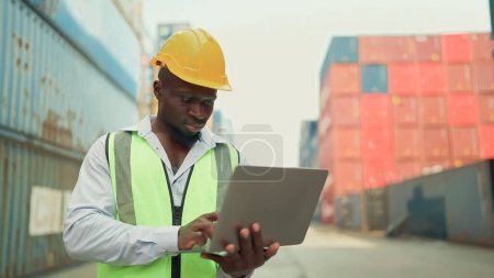 Photo for African American Black industrial engineer in yellow hard hat and safety vest standing, working on laptop checking container at container yard warehouse. Shipping import export, Logistics business - Royalty Free Image