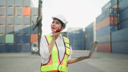Téléchargez les photos : Ingénieur asiatique femme en uniforme de sécurité parler sur talkie-walkie et woking sur ordinateur portable pour le contrôle de chargement de conteneurs boîte de fret du navire de fret pour l'exportation d'importation. Logistique, Concept de transport - en image libre de droit