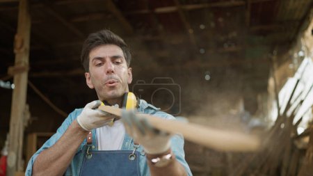 Photo for Carpenter blowing sawdust from wooden plank. Young carpenter man blowing sawdust from wooden detail while working in carpentry workshop. Carpenter and woodcraft concept - Royalty Free Image
