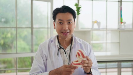 Foto de Consulta médica en línea. Médico dentista masculino asiático que muestra modelo de mandíbula educar sobre el cuidado de las encías y los dientes a través de hablar en línea, haciendo videollamada con el paciente. Medicina y tecnología - Imagen libre de derechos