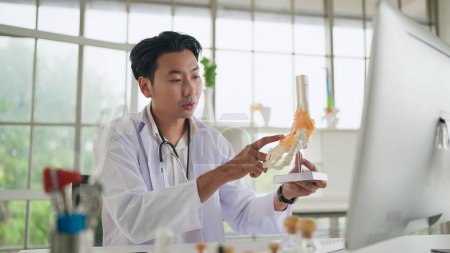 Téléchargez les photos : Consultation médicale en ligne. Médecin orthopédique masculin asiatique montrant le squelette du pied, modèle d'anatomie articulaire de la cheville expliquer pour le patient en parlant en ligne, faire un appel vidéo. traitement de physiothérapie - en image libre de droit