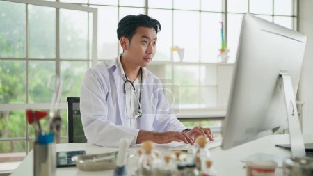 Téléchargez les photos : Jeune asiatique homme médecin à l'aide d'un ordinateur travaillant à la clinique ou à l'hôpital. Docteur tapant sur ordinateur bavarder en ligne avec le patient pour aider la consultation, expliquer la maladie. Médecine et technologie - en image libre de droit