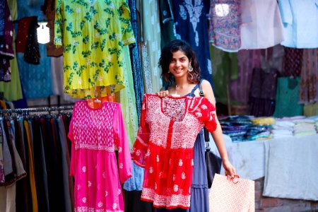 Foto de Hermosa mujer sexy mostrando corto color rojo chikankari bordado a mano kurti en la mano posando al aire libre en el mercado callejero mientras mira a la cámara - Imagen libre de derechos