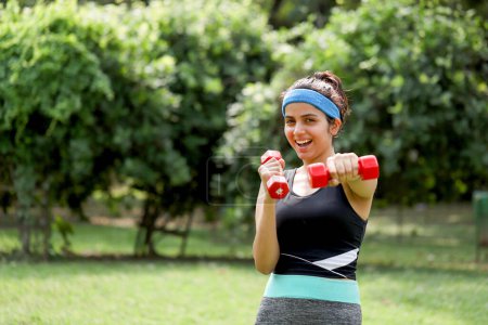 Photo for Hot fit woman posing with dumbbells and happily smiling at the camera while standing in the park - Royalty Free Image