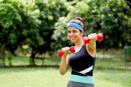 Sexy athletic woman in gym outfit smiling while holding dumbbell exercise in park