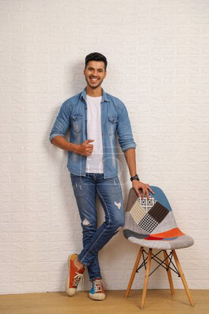 Smiling nice man standing next to a chair wearing a denim outfit and posing towards the camera