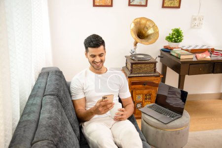Photo for Sexiest man sitting on sofa in living area smiling while watching reel on social media on mobile phone - Royalty Free Image