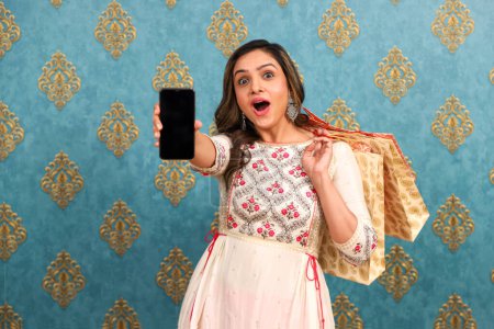 Una hermosa joven dama de traje tradicional durante el festival Diwali está sosteniendo bolsas de compras y mostrando la pantalla de su teléfono a la cámara