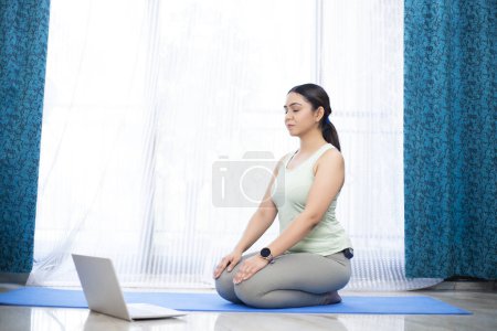 Photo for Peaceful woman meditating in front of laptop at home, sitting in vajrasana and doing breathing exercises - Royalty Free Image