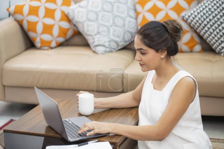 Mujer madura sentada en el suelo en la sala de estar y trabajando en el ordenador portátil bebiendo café