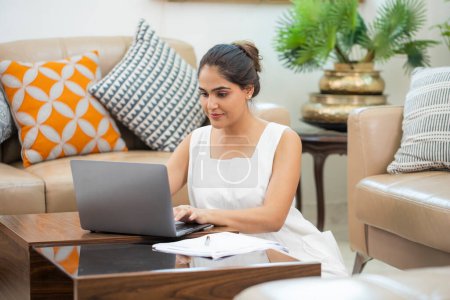 Frau sitzt in weißem Freizeitkleid auf dem Boden und arbeitet zu Hause am Laptop