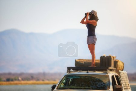 Photo for On safari in Zambia: Silhouette of a Fit Woman standing on the roof of safari car, observing Zambezi River Nature through binoculars. Lower Zambezi National Park, Zambia, Africa. - Royalty Free Image