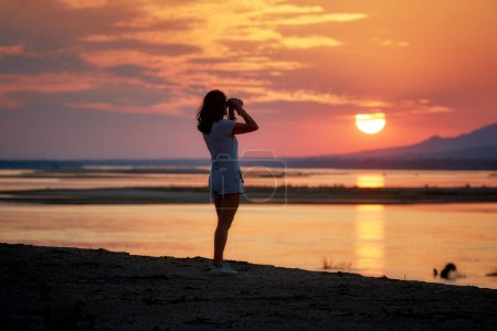 Photo for On safari in Zambia: Silhouette of a Fit Woman standing on the bank of  Zambezi River against sunset, observing  Nature through binoculars. Orange and red colors, Lower Zambezi National Park, Zambia, Africa. - Royalty Free Image