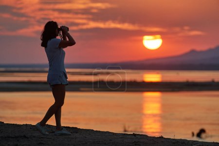 Foto de On safari in Zambia: Silhouette of a Fit Woman standing on the bank of Zambezi River against sunset, observing Nature through binoculars (en inglés). Colores naranja y rojo, Parque Nacional Lower Zambezi, Zambia, África. - Imagen libre de derechos