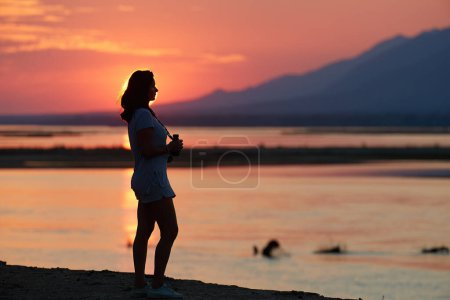 Photo for On safari in Zambia: Silhouette of a Fit Woman standing on the bank of  Zambezi River against sunset, observing  Nature through binoculars. Orange and red colors, Lower Zambezi National Park, Zambia, Africa. - Royalty Free Image