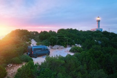 Photo for Camper van stands close to shining lighthouse on rocky hill with forest against epic pink sunset. Wanderlust lifestyle, travel adventures in Cape Ducato or Doukato, Lefkada, Greece. - Royalty Free Image