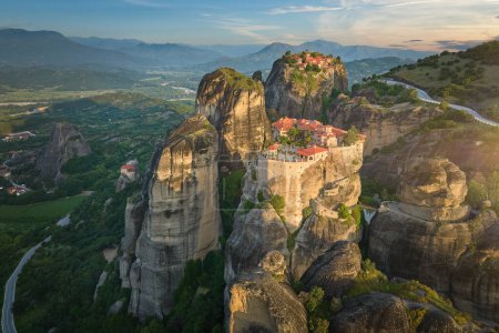 Aerial view of Meteora Monastery complex on rocks, illuminated by rising sun against mountains in background. UNESCO World Heritage Site. Greece.