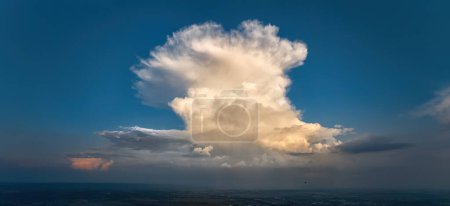 Photo for Panoramic aerial shot of cumulonimbus capillatus, a huge storm cloud cumulonimbus with dense cirrus clouds above against a blue sky and the silhouette of a small hot air balloon. - Royalty Free Image