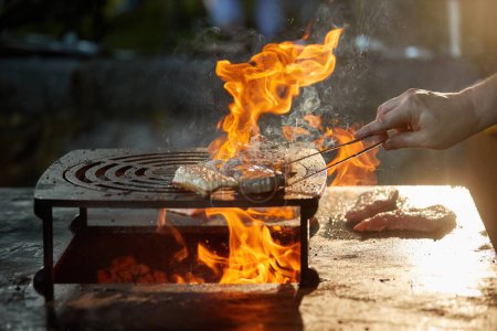 Téléchargez les photos : Un chef prépare un steak sur un gril extérieur. Gros plan sur la cuisine au festival du jardin, flammes, faible profondeur de champ, fond flou très coloré, soirée au festival du jardin. - en image libre de droit