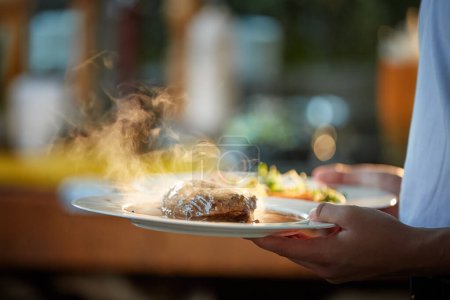 Photo for Serving food at the Garden Party.    Two plates in hand, with steak and salmon, in coloured, blurred backlight, shallow depth of field, backlit steam rising from the food. - Royalty Free Image